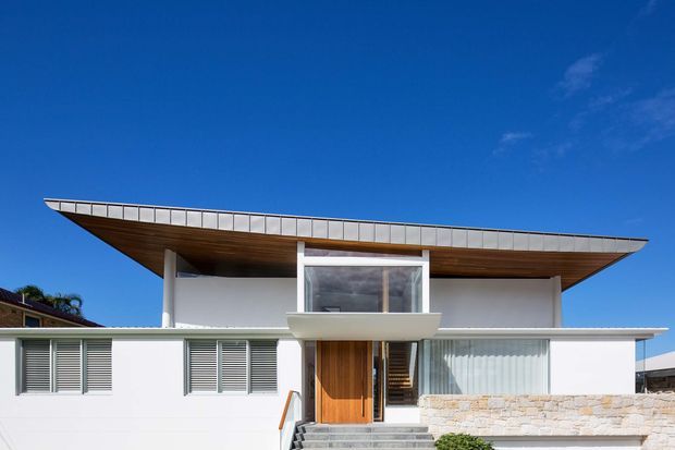 Front exterior view of a modern two-storey residence with a broad projecting low-pitched roof, white rendered walls, extensive glazing, timber soffit, timber entry door, horizontal louvred shutters, and a stone-clad lower facade. The home is photographed against a clear blue sky.