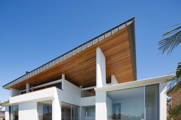 Exterior view of a contemporary multi-level residence under a clear blue sky. The design features a dramatic low-pitched roof with a deep timber-lined soffit, white rendered structural walls and columns, extensive glazing, and a glazed upper-level balustrade. Palm foliage is visible at the edges of the site.