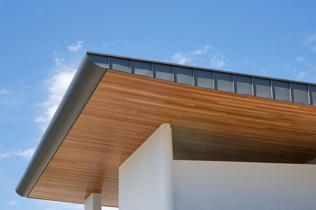 Close-up upward view of a modern building facade with a broad projecting roof or canopy. The soffit is clad in warm horizontal timber, supported by smooth white structural walls and columns, with a continuous band of dark-framed clerestory glazing along the roof edge. The image has a clean contemporary architectural character against a blue sky.