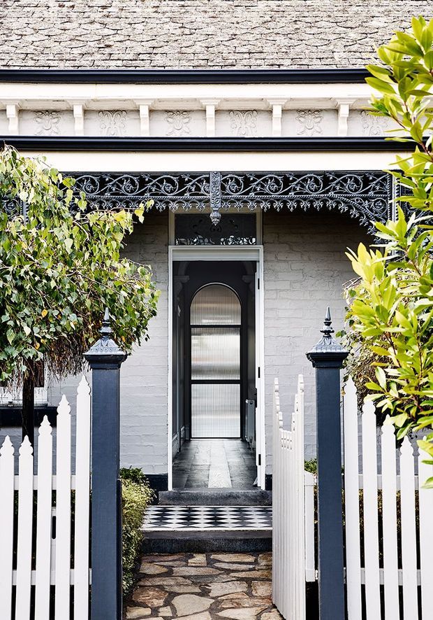 Front elevation of a heritage-style residence viewed through an open white picket gate. The pale painted masonry facade features ornate lacework verandah detailing, decorative frieze panels, dark trim, a glazed entry door, patterned black-and-white tiled threshold, stone pathway, and richly detailed entrance posts framed by garden planting.