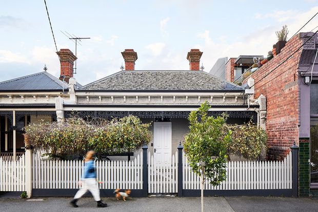 Street-facing view of a single-storey heritage terrace or cottage with a low hipped slate roof, multiple brick chimneys, ornate Victorian-era lacework and verandah detailing, painted masonry, and a white picket fence. A contemporary brick building is visible at the right edge. The image is an exterior architectural streetscape.