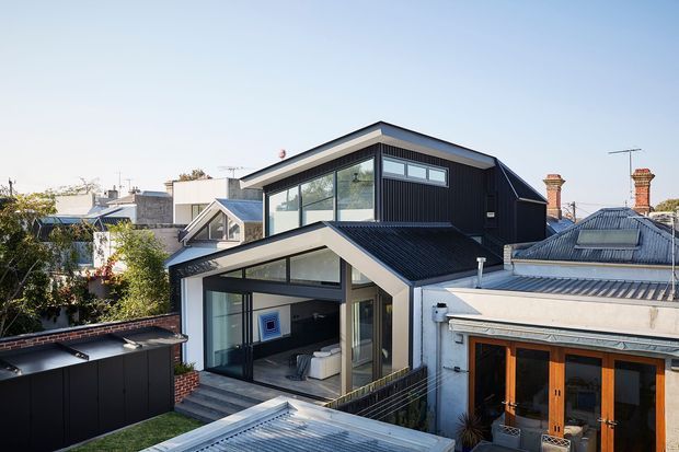 Exterior view of a contemporary multi-level home extension in a dense suburban setting. The dark vertically clad upper volume has a pitched gable roof, clerestory-style glazing and extensive windows, while the lower level features a glazed living area, covered terrace and dark metal roofing. Adjacent older roof forms and brick chimneys remain visible, creating a contrast between the new and existing structures.