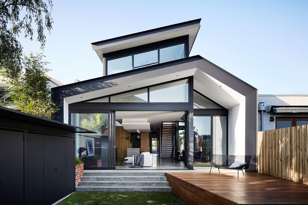 Exterior view of a contemporary multi-level residence with a bold asymmetrical pitched roof, extensive floor-to-ceiling glazing, dark window frames, and a white rendered facade. The image shows a timber deck, concrete entry steps, a dark garage or carport structure, and a glimpse of the open-plan interior with a staircase through the glazing.