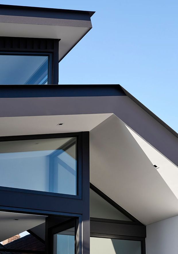 Upward-looking architectural photograph of a sharply composed modern house exterior, featuring intersecting flat and pitched roof planes, deep eaves, white soffits, dark charcoal cladding and large black-framed glazed windows against a clear blue sky.