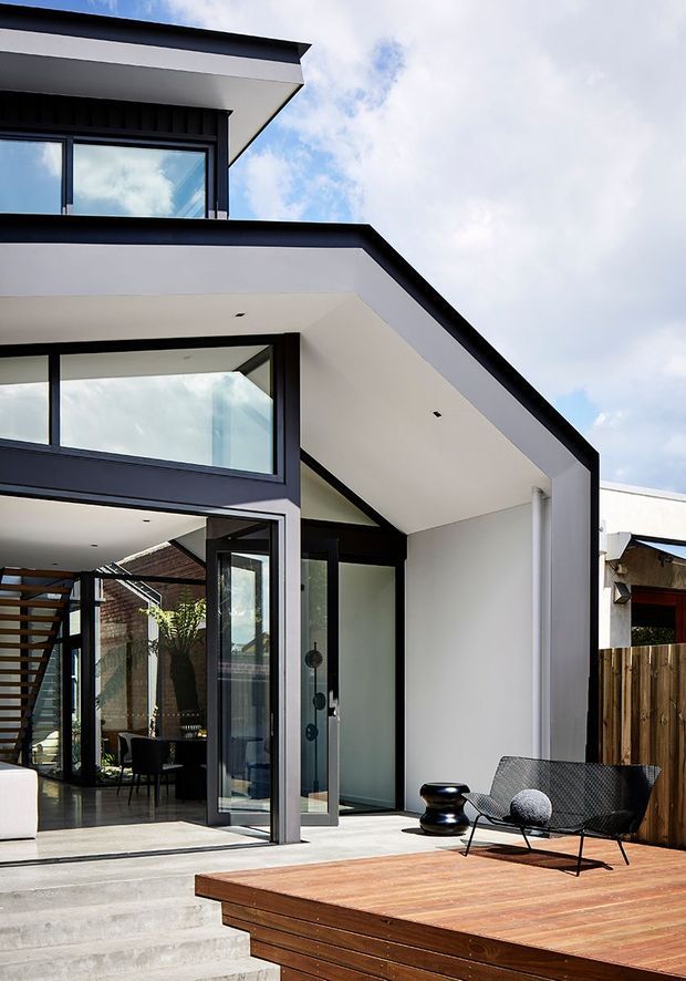 A contemporary multi-level residential extension viewed from an outdoor deck. The white rendered structure has bold black trim, projecting flat roof planes, large dark-framed glazed openings, and a partially visible interior dining area. Broad concrete steps lead up to a warm timber deck furnished with a modern outdoor lounge chair and low sculptural table. A timber fence and neighbouring rooflines are visible in the background beneath a partly cloudy sky.