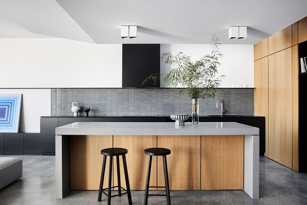 A contemporary open-plan kitchen dominated by a large island with a light stone benchtop, vertical timber cabinetry, and two black stools. The room features a dark grey horizontal tile splashback, black lower cabinetry and appliances, white walls and ceiling, polished concrete flooring, integrated timber wall storage, and minimalist square ceiling lights. A leafy indoor tree provides a natural focal point.
