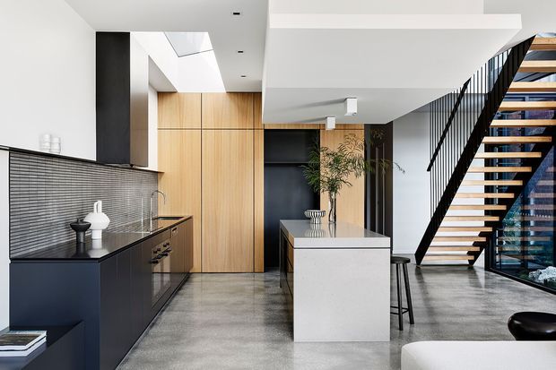A bright, open-plan contemporary kitchen beneath a stairway. The space features dark matte cabinetry, warm timber wall cabinetry, a large pale island, a dark textured tiled splashback, polished grey concrete flooring, and an open timber-tread staircase with black metal balustrades. Floor-to-ceiling glazing and clean white ceilings create an airy, refined interior.