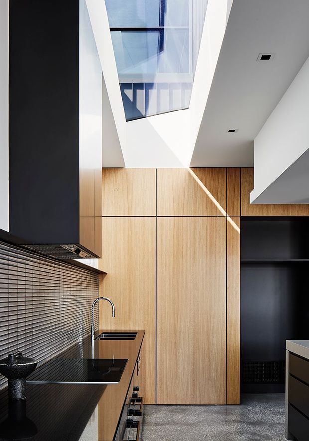 A compact contemporary kitchen with tall light-wood cabinetry, a black stone-look benchtop, dark tiled backsplash, integrated black appliances, and a dramatic overhead skylight. Strong geometric lines and high-contrast finishes create a sleek, refined interior.