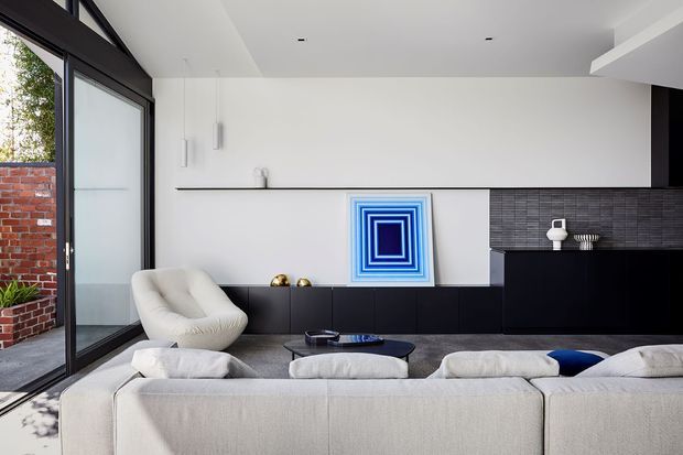Contemporary living room viewed from behind a large pale sectional sofa, with full-height black-framed glazing opening to a brick courtyard. The room features a white wall with minimal floating cabinetry, a black built-in media or storage wall, dark textured tile around a fireplace or feature zone, recessed ceiling lights, and vivid blue geometric artwork. The palette is monochromatic with a strong blue accent and a refined, minimalist character.