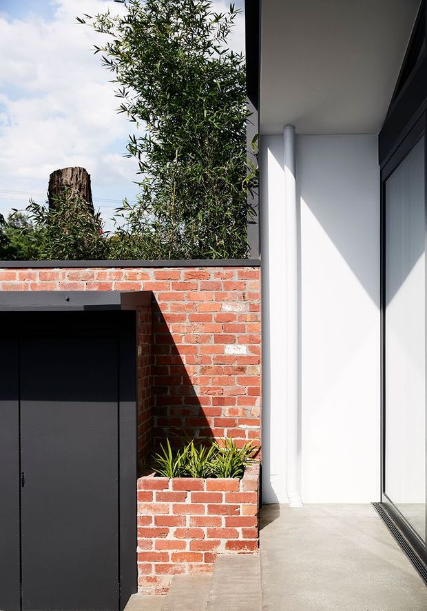 A close architectural view of a contemporary residential exterior courtyard or entry area. A red brick wall incorporates a raised planter and frames a dark matte garage or storage door, while a crisp white rendered volume with a downpipe and large dark-framed glazed opening sits to the right. Concrete paving, strong sunlight, and established greenery create a contrast between heritage-style brickwork and modern additions.