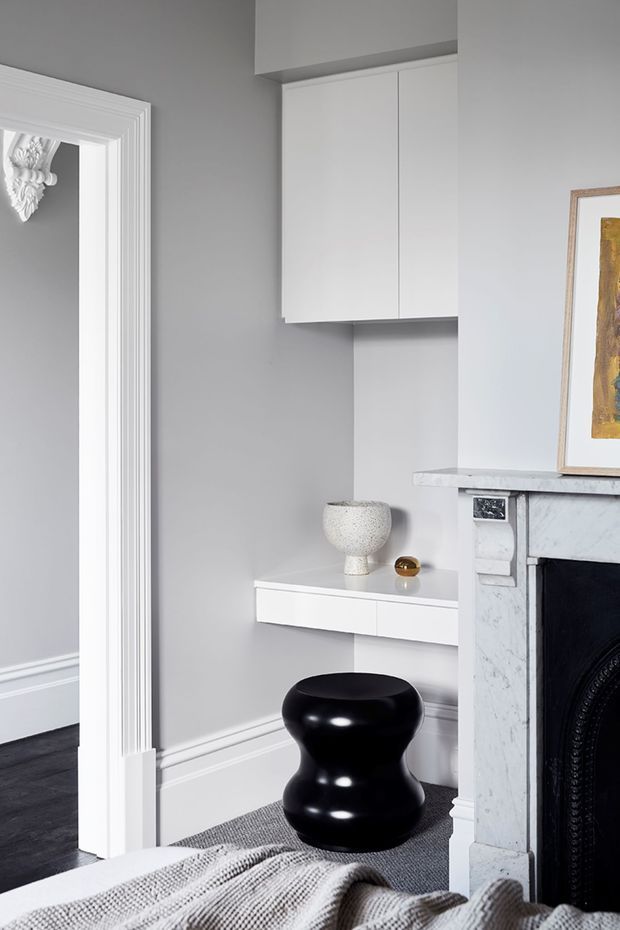 A refined grey-and-white interior corner featuring a classic marble fireplace with ornate white trim, a floating white built-in console and overhead cabinetry, and a glossy black sculptural stool. Decorative objects and framed artwork add restrained accents, while a doorway with detailed moulding opens to another room.