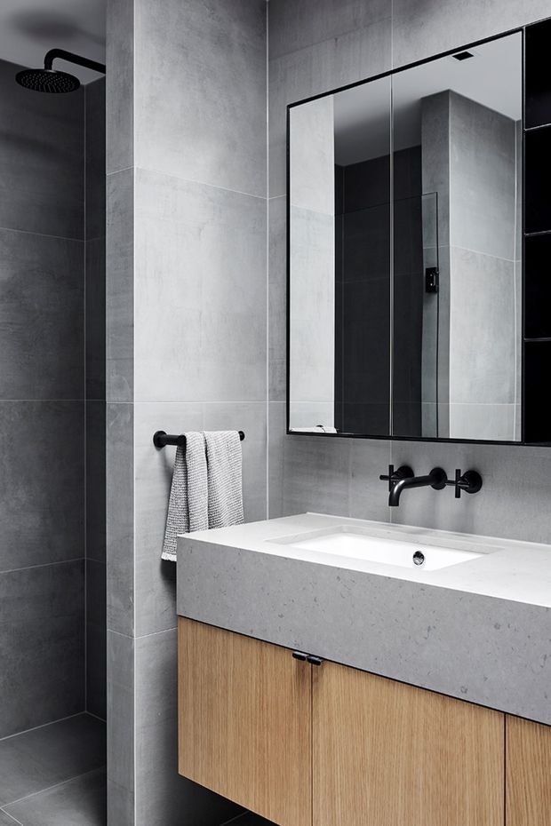 A contemporary bathroom featuring large-format grey concrete-look wall and floor tiles, a frameless glass shower with matte black rainfall showerhead, a wall-mounted black tap set, a broad white integrated basin and benchtop, and a light timber floating vanity. A large mirrored cabinet sits above the vanity, with a simple towel rail beside the shower.