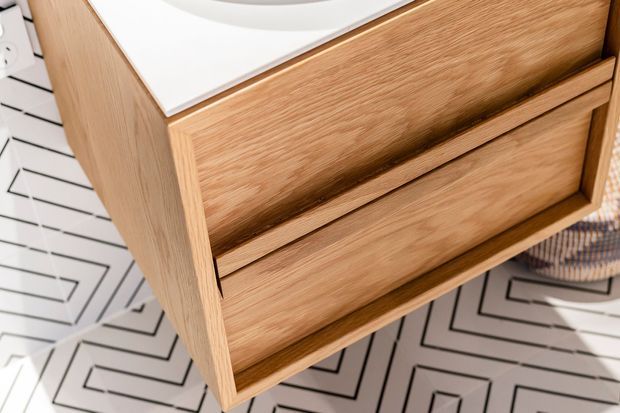 Close-up of a wall-mounted or compact bathroom vanity in natural light oak timber, featuring two recessed-front drawers and a white countertop with an inset basin. The vanity sits above a patterned geometric floor, with white geometric wall tiling visible at the left edge.