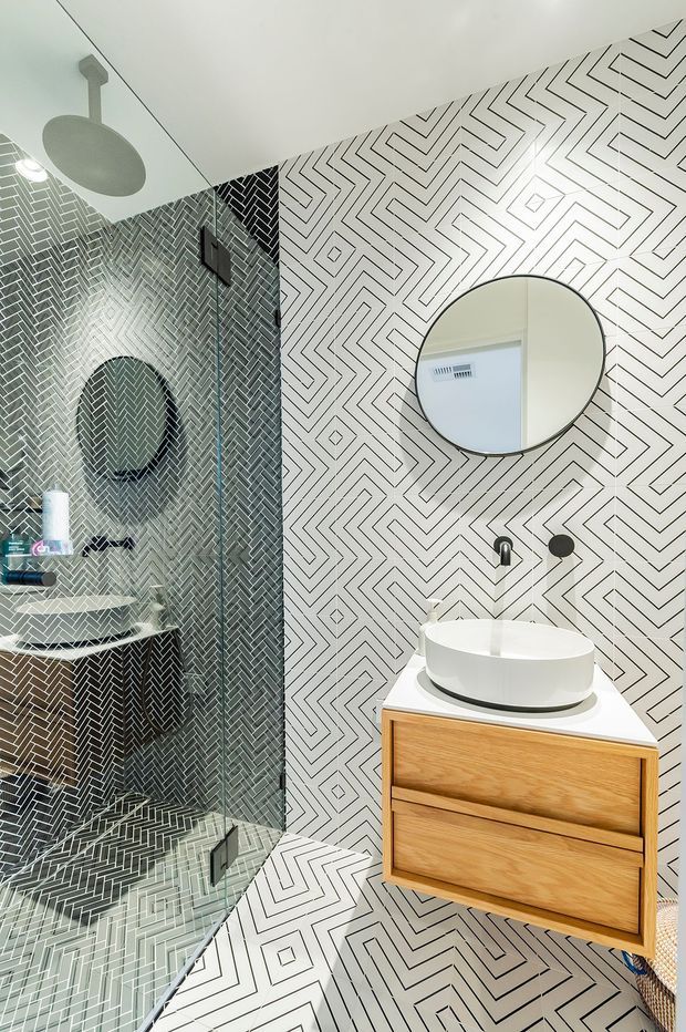 A compact contemporary bathroom featuring bold black-and-white geometric patterned tiles across the walls and floor. The room includes a frameless glass shower enclosure with a round ceiling showerhead, a round wall mirror, matte dark fixtures, and a floating warm timber vanity with a white vessel basin. The high-contrast graphic tile pattern creates a striking, playful focal point.