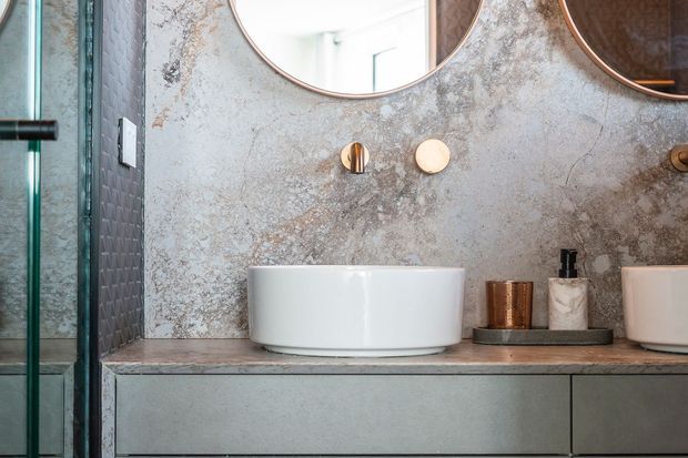 Close-up of a contemporary bathroom vanity with a white vessel basin set on a stone-look benchtop, integrated grey cabinetry below, textured grey wall finish, round copper-framed mirrors, and warm metallic tapware and accessories. A glass shower enclosure is visible at the left edge.