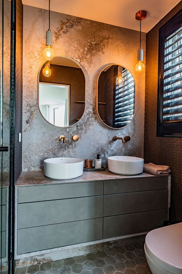 A moody contemporary bathroom featuring a double floating vanity with two white vessel basins, brushed brass wall-mounted tapware, rounded rectangular mirrors, and warm pendant lights. The walls have a mottled concrete-look finish, the vanity is in dark grey-green cabinetry, and the floor is covered in dark hexagonal tiles. A toilet, textured dark wall finish, and louvred window are visible on the right.