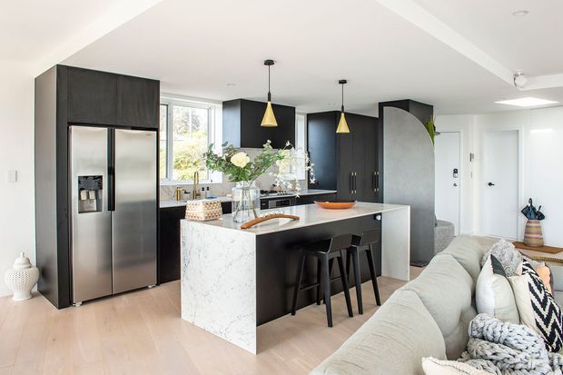 Open-plan contemporary kitchen and living area with a large marble-look island, dark matte cabinetry, stainless-steel refrigerator, black bar stools, pendant lights, light timber flooring, and a pale sectional sofa. Bright natural light enters through the rear glazing, with a hallway and doors visible beyond the kitchen.