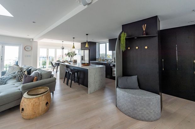 Open-plan contemporary interior combining a living room and kitchen. The space features pale timber flooring, a large light-grey sectional sofa, a dark kitchen island with a pale veined stone benchtop, matte-black built-in cabinetry and feature wall, pendant lights, and generous glazing with views outside. A rounded upholstered occasional chair and timber side table add softer organic forms.