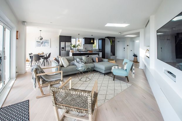 Bright open-plan living area with a contemporary light neutral interior. The foreground contains two woven timber rocking chairs, while a large pale sectional sofa, patterned cushions, coffee tables, and a pale geometric rug define the living zone. A dining table sits behind the sofa, with a dark fixed kitchen island and cabinetry beyond. Full-height glazing is visible on the left, and a wall-mounted television with built-in white media joinery occupies the right wall.