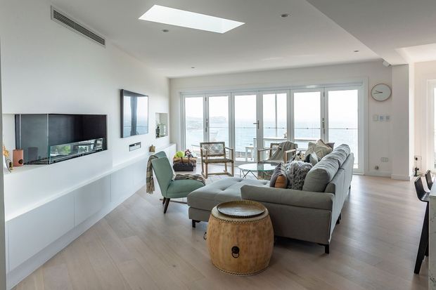 Bright contemporary living room with pale timber flooring, white walls, a built-in media wall, large sectional sofa, armchairs and a glass coffee table. Full-height glazed doors open to a balcony or terrace with a visible waterfront beyond, while a skylight adds natural light.