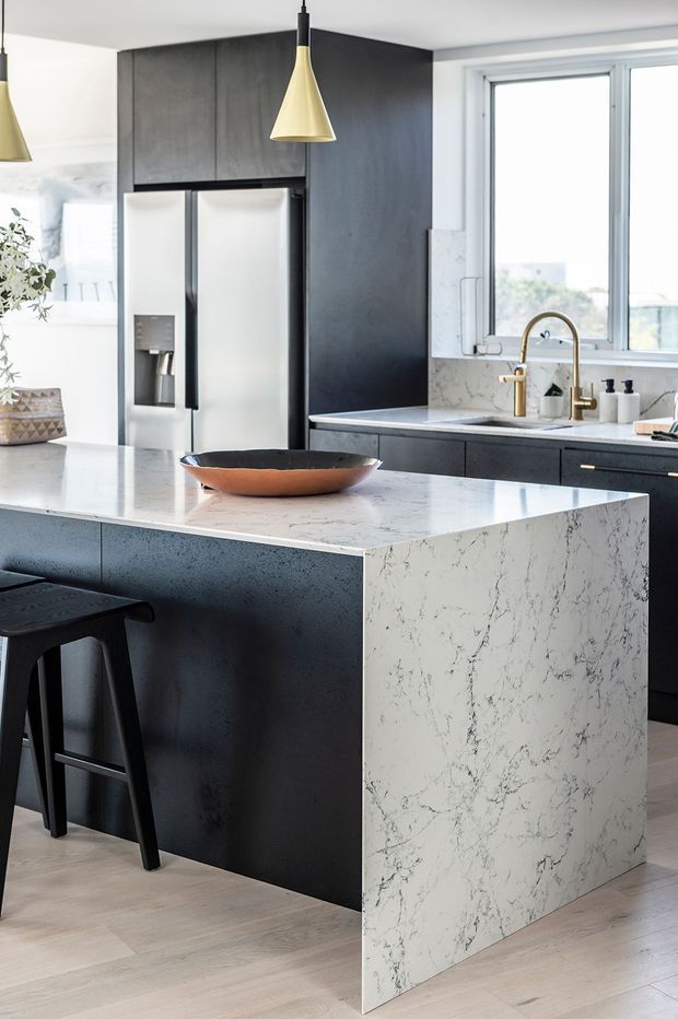 Contemporary kitchen centered on a large island with a white veined stone waterfall benchtop and dark charcoal cabinetry. The space includes a black double-door refrigerator, black bar stools, a white marble-look splashback, gold gooseneck tapware, and two gold-and-black pendant lights. Large windows provide abundant natural light, while pale timber flooring adds warmth.