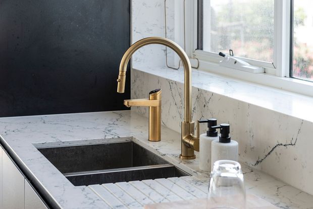 Close-up of a contemporary kitchen sink area beneath a window, featuring a white veined stone benchtop and splashback, a dark undermount double-bowl sink, and coordinated brushed brass gooseneck and secondary tapware. White-framed glazing and a dark charcoal wall create contrast, with soap dispensers and a draining rack on the bench.