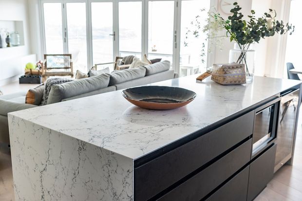 Bright open-plan kitchen with a large white veined stone island featuring dark built-in drawers and integrated appliances. A light-filled living area with a grey sectional sofa and floor-to-ceiling glazed doors is visible behind the island, with coastal-looking views beyond.