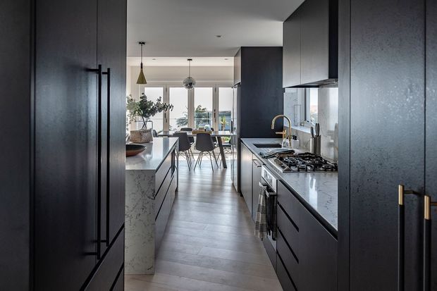 A dark, sophisticated galley-style kitchen viewed toward an open dining area. Tall charcoal cabinetry frames the image, with a central marble-look island, matching stone benchtops, black lower cabinetry, brass tapware and handles, integrated appliances, and a gas cooktop. Beyond the kitchen is a dining table with pendant lighting and large windows providing natural light.