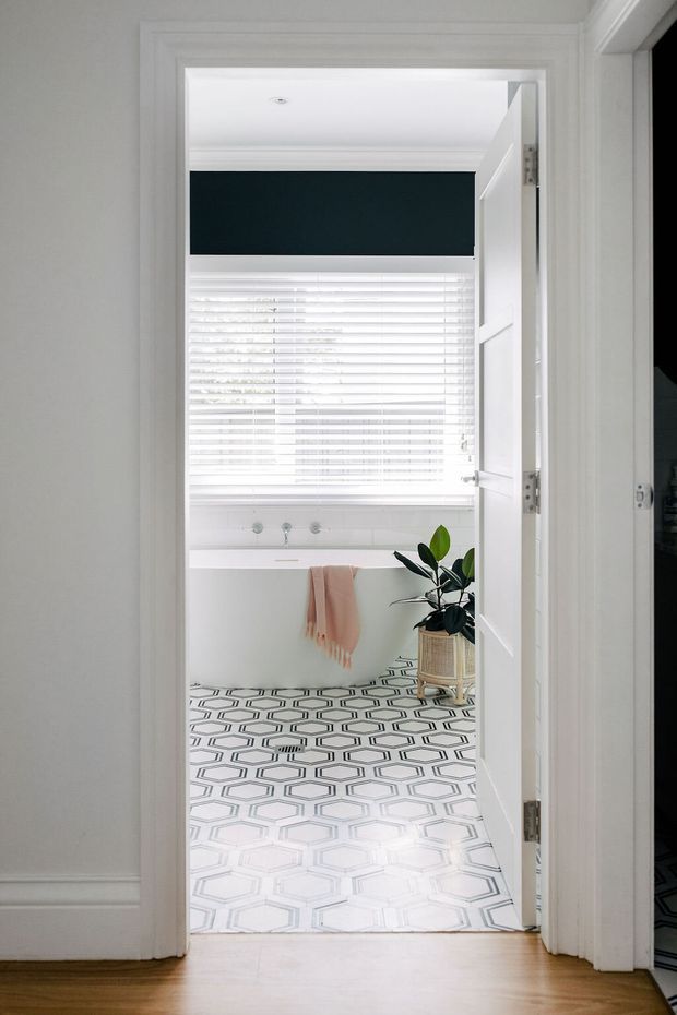 View through an open white paneled door into a bright bathroom. The room features a patterned geometric tile floor, white cabinetry and wall finishes, a wide window with white plantation blinds and a dark teal valance, a towel rail with a blush towel, and a potted plant. Timber flooring is visible in the hallway outside.