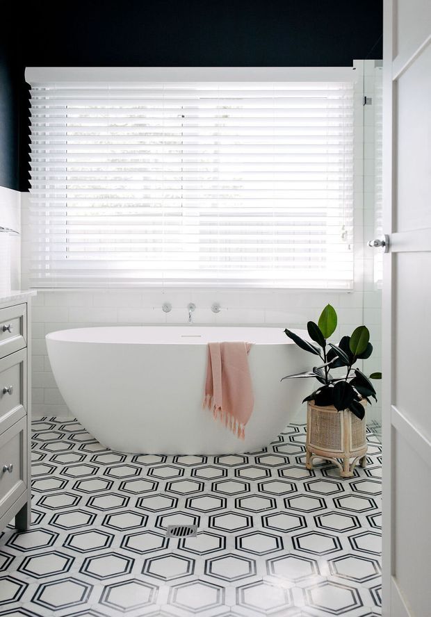 Bright bathroom featuring a freestanding white bathtub beneath a wide window with white blinds, white tiled walls, and a bold black-and-white geometric tiled floor. A pale pink towel hangs over the tub, while a leafy potted plant and white vanity add softness. The room has a clean, high-contrast, contemporary look.