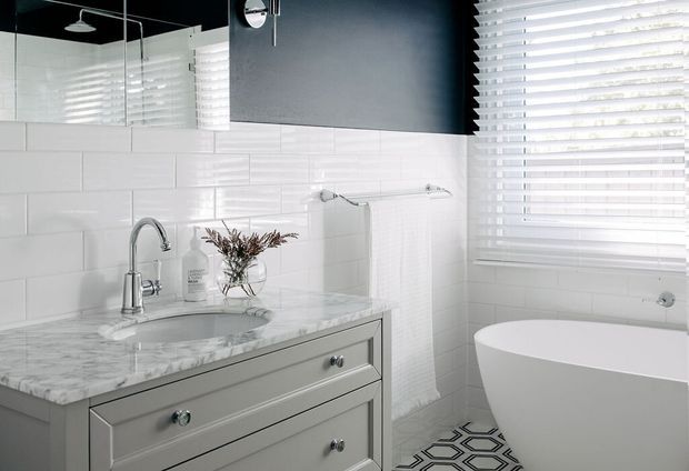 Bright contemporary bathroom featuring a grey double-drawer vanity with a white marble-look benchtop, chrome tapware, glossy white horizontal subway wall tiles, a freestanding white bathtub, white venetian blinds, and a bold dark geometric floor tile pattern. A large mirror reflects the shower area.