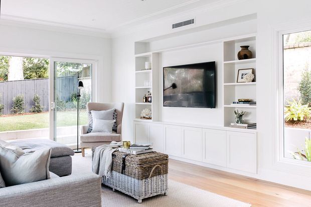 Bright, refined living room with white built-in entertainment cabinetry and open display shelving surrounding a wall-mounted television. The room includes a large neutral sectional sofa, upholstered armchair, woven storage ottoman, pale timber flooring, a soft area rug, expansive windows, and sliding glass doors opening to a fenced garden. The overall look is light, comfortable, and contemporary with classic trim detailing.