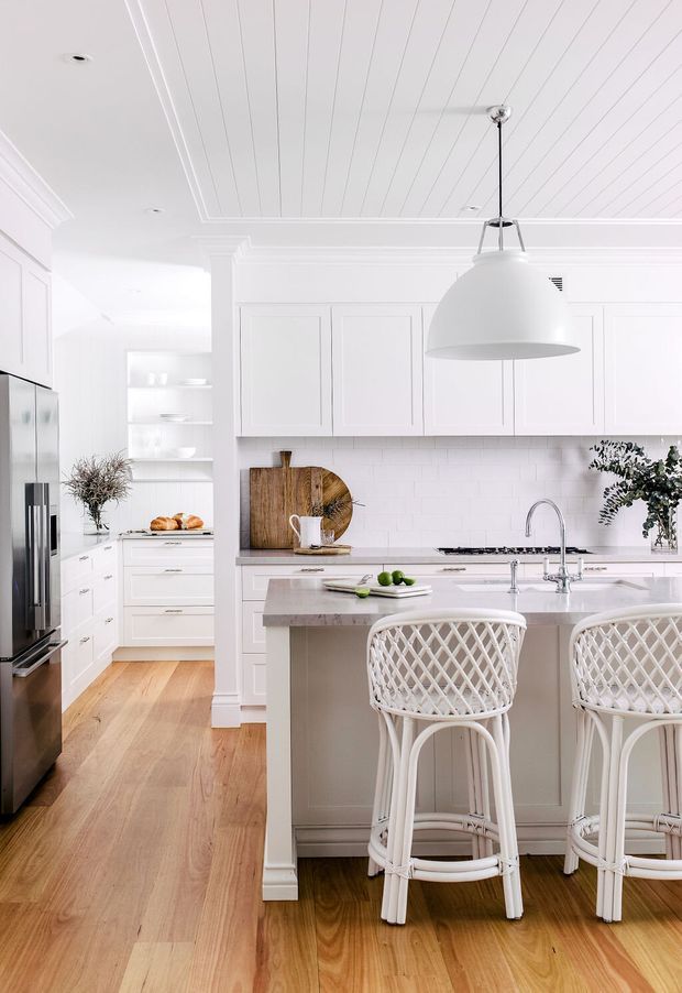 Bright, refined white kitchen with a central island and two woven-back bar stools. The room features white shaker-style cabinetry, a white subway-tile splashback, pale stone benchtops, warm timber flooring, integrated stainless-steel appliances, open shelving, and a large white pendant light. Decorative greenery and timber boards add subtle natural accents.