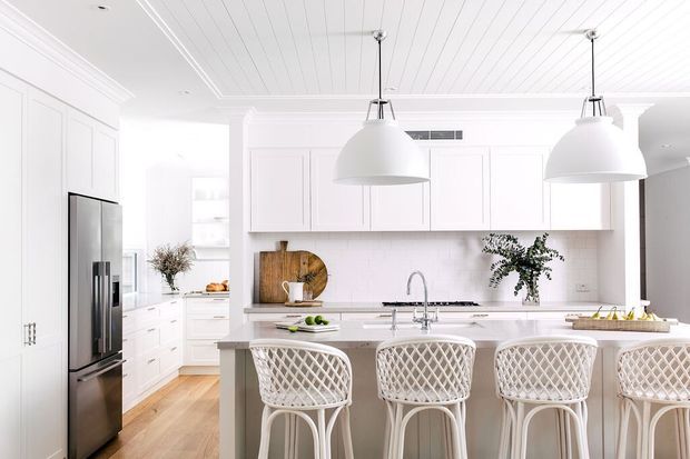 Bright white kitchen with a large central island, four woven bar stools, shaker-style cabinetry, stainless-steel refrigerator, pale timber flooring, white panelled ceiling, and two oversized pendant lights. The space has a clean, refined coastal character with abundant natural light.