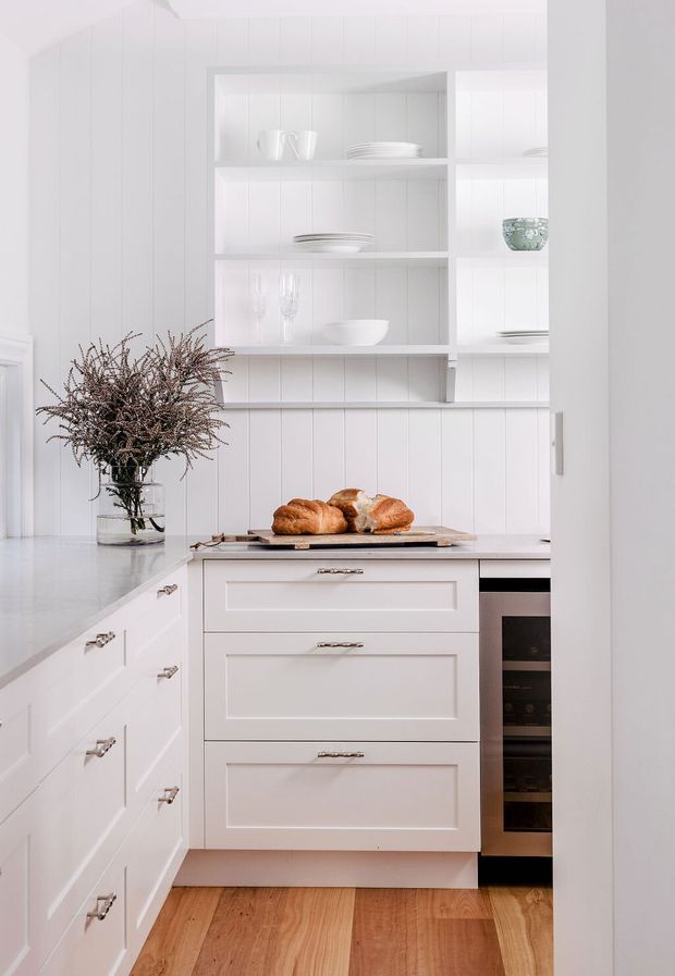 Bright, compact white kitchen with shaker-style cabinetry, brushed metal pulls, a pale benchtop, vertical white wall panelling, and open display shelving holding crockery. A timber chopping board with bread sits on the benchtop, while warm timber flooring contrasts with the predominantly white fit-out. A built-in stainless-steel oven is integrated beside the drawers, and a vase of branches adds a natural accent.