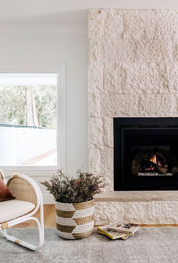 Bright, refined living room corner featuring a large textured light stone fireplace with a black firebox, a white-trimmed window, pale timber flooring, and a woven accent chair beside a striped woven planter. A soft grey rug, books, and restrained natural styling create a calm contemporary-traditional atmosphere.