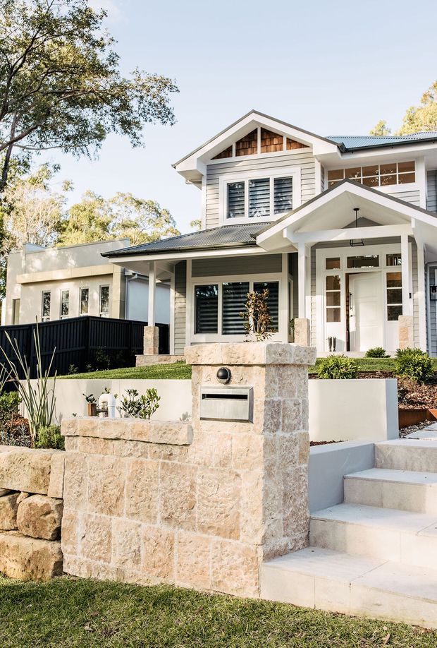 A bright street-facing view of a two-storey suburban home with a gabled roof, white weatherboard cladding, dark metal roofing, and a covered entry porch. The facade includes white-framed windows with shutters and a timber-lined gable detail. Pale natural-stone retaining walls and a stone mailbox pillar frame the landscaped front garden and broad entry steps.
