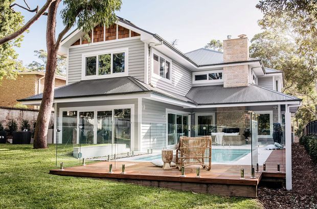Exterior rear view of a large two-storey suburban home with light horizontal weatherboard cladding, dark corrugated metal roofing, broad glazed openings, and a prominent pale stone chimney. A timber deck with frameless glass balustrade overlooks a lawn, while a glazed lower-level living area and pool or water feature are visible behind it. Mature trees surround the property.