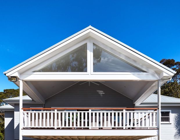 Exterior front view of a light-coloured gable-roofed house with a prominent covered upper-level balcony or verandah. The facade features a large triangular glazed gable, white timber trim and balustrading, grey horizontal weatherboard cladding, and a corrugated metal roof. The image is photographed in bright daylight against a clear blue sky.