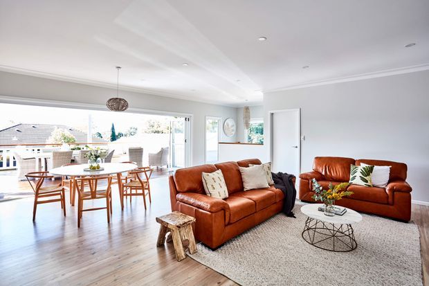 Bright open-plan living and dining area with warm brown leather sofas, a timber dining table and chairs, pale wood flooring, a large textured rug, and expansive glazed doors opening to an outdoor balcony or terrace. The room has white walls and ceilings with simple cornice detailing, recessed downlights, and a woven pendant above the dining table.