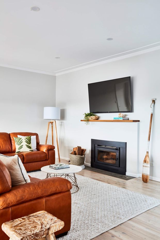 Bright, refined living room with white walls and decorative ceiling cornice, pale timber flooring, a black built-in fireplace, wall-mounted television, and a simple timber mantel shelf. The room is furnished with cognac leather sofas, a white round coffee table, a woven light rug, a tripod floor lamp, a firewood basket, and coastal-inspired paddle decor.