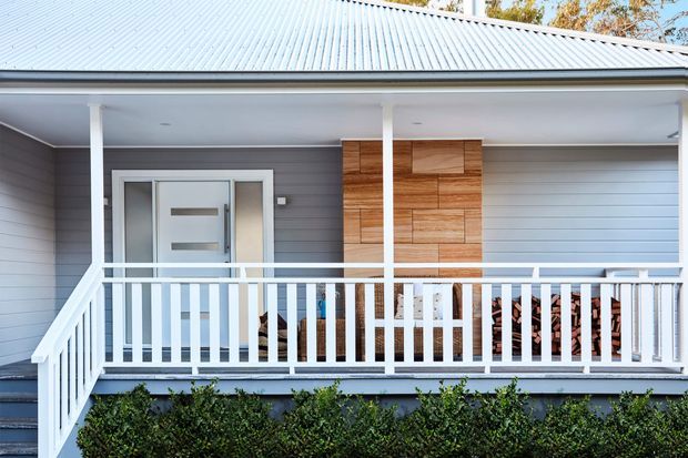 Front exterior of a contemporary single-storey home with a covered entry porch, pale grey horizontal weatherboard cladding, a light grey metal gabled roof, white posts and balustrades, a modern glazed entry door, and a warm timber-clad feature wall beside the garage. Landscaping is visible along the base.