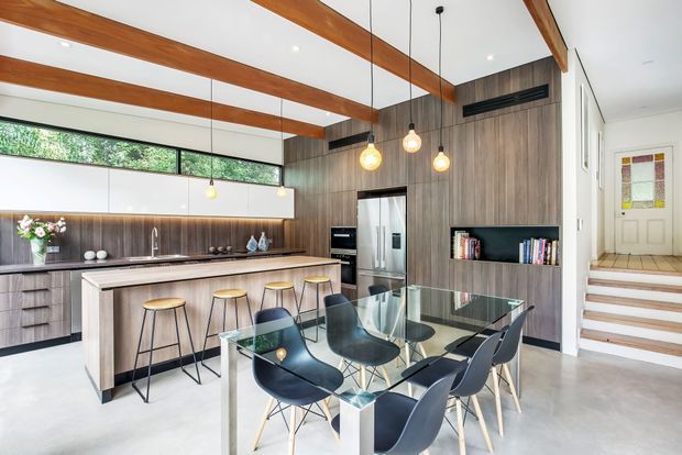 Bright open-plan kitchen and dining area with a contemporary timber-and-white fit-out. The kitchen features a long island with integrated sink and seating, dark timber-look cabinetry, stainless-steel appliances, and a white splashback. Exposed warm timber ceiling beams and pendant lights span the space. A glass dining table with dark molded chairs sits in the foreground, while stairs lead to an entry area at right.