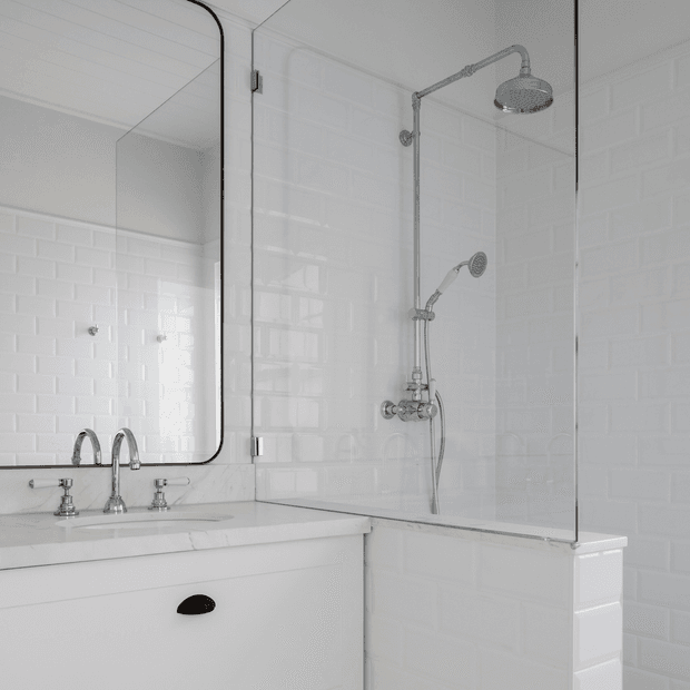 Bright, refined bathroom with a white vanity, marble-look benchtop, chrome tapware, large black-trimmed mirror, and a glass-enclosed shower featuring both overhead and handheld fixtures. White subway wall tiles create a clean, classic backdrop.