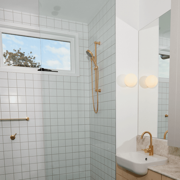 Bright, compact bathroom with pale blue-grey square wall tiles, a frameless glass shower enclosure, white-framed window, and brushed brass shower and tapware. A wall-mounted white basin sits on a timber vanity with a stone-look benchtop, beside a large mirrored cabinet and globe wall lights.
