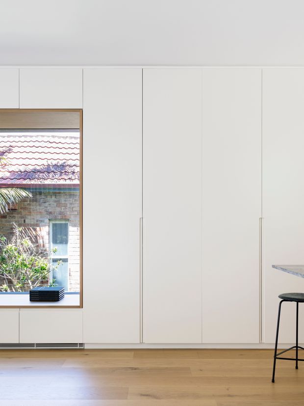 Bright minimalist interior featuring a full-height wall of flush white built-in cabinetry with slim vertical handles, a warm light timber floor, and a timber-framed window overlooking a brick exterior courtyard. A small portion of a stone benchtop and a black bar stool are visible at the right edge, suggesting a kitchen or adjoining kitchen area.