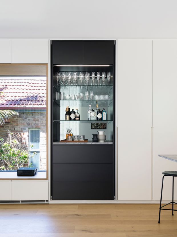 A contemporary built-in drinks bar integrated into full-height white cabinetry. The dark charcoal joinery has illuminated glass shelves displaying wine glasses, tumblers, bottles and barware, with drawers below and a pale stone countertop. A timber-framed window and light timber flooring add warmth, while a partial kitchen island and stool are visible at right.
