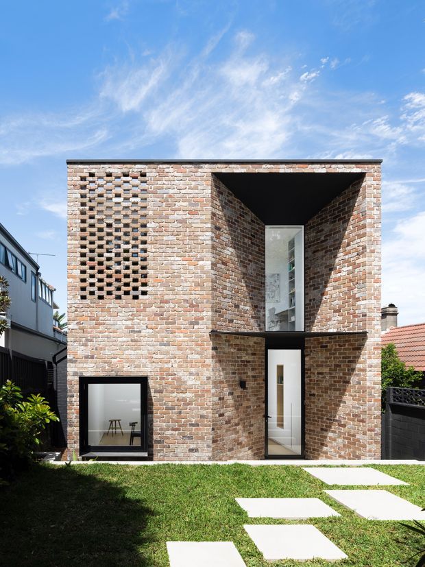 Exterior view of a compact two-storey brick residence with a strongly geometric composition. The facade combines warm recycled-looking brickwork, a recessed upper-level void, a projecting dark horizontal balcony or canopy, a perforated brick screen, and tall narrow glazing. A small lawn with concrete stepping slabs leads toward the house, with neighbouring urban homes visible at the sides.