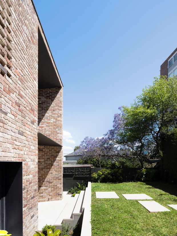 Exterior architectural photograph of a contemporary brick building beside a landscaped residential courtyard. The tall exposed brick wall has deep recessed openings and strong geometric shadows, with pale concrete steps and paving leading into a grassed garden. Mature trees and neighbouring buildings are visible beyond the courtyard under a clear blue sky.