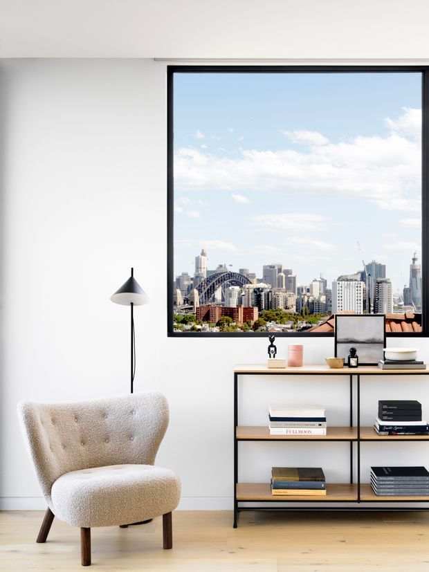 Bright, minimalist living room with white walls, pale timber flooring, a sculptural boucle armchair, slim black floor lamp, and an open black metal shelving console styled with books and small decor. A large black-framed picture window overlooks a dense city skyline with a prominent arched bridge.